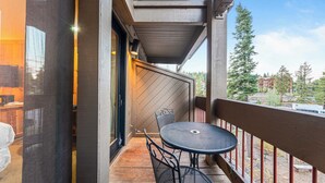 A balcony with a round table and two chairs, a wooden deck, and a view of a mountainous area with buildings. - Cozy Northstar Village Escape w/ Forest Views & Easy Lift Access Alpineglow Vista by Avantstay (Truckee)