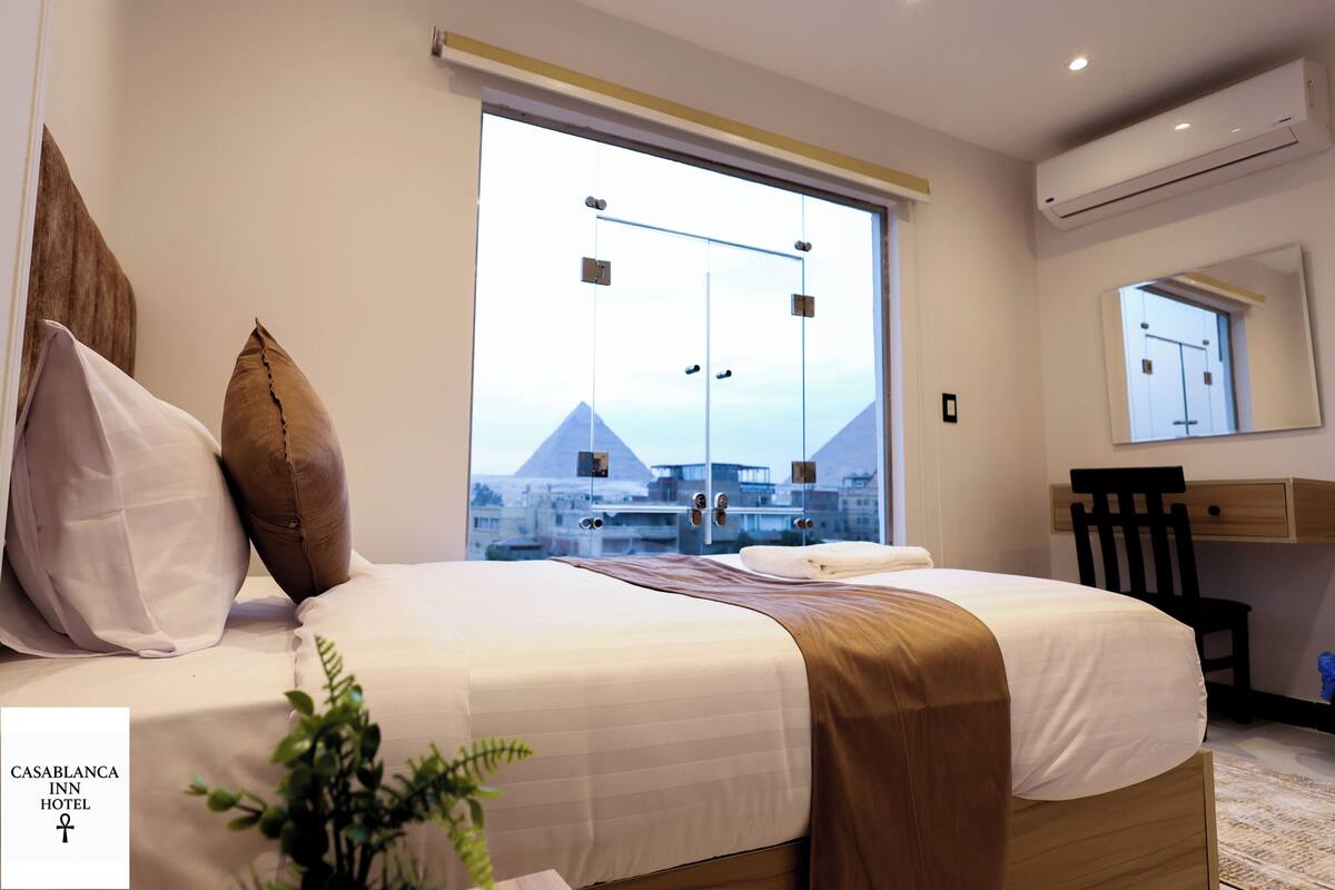 A hotel room with a bed, a desk, a chair, and a large window with a view of buildings.