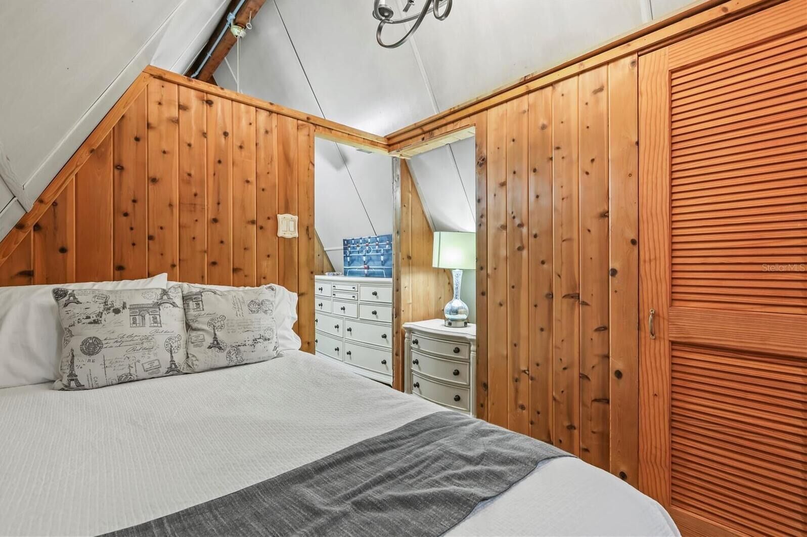 Une chambre avec un plafond en pente, des murs recouverts de lambris en bois, un lit agrémenté d’oreillers à motifs, une commode et une lampe.