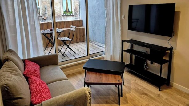 En opholdsstue med sofa, et lille bord, et fjernsyn og en skydedør til et udendørs område med et bord og stole.