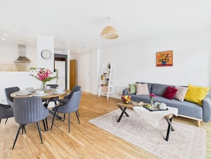 A modern living room with a dining area, a grey sofa with colorful cushions, a wooden coffee table, and a white bookshelf.