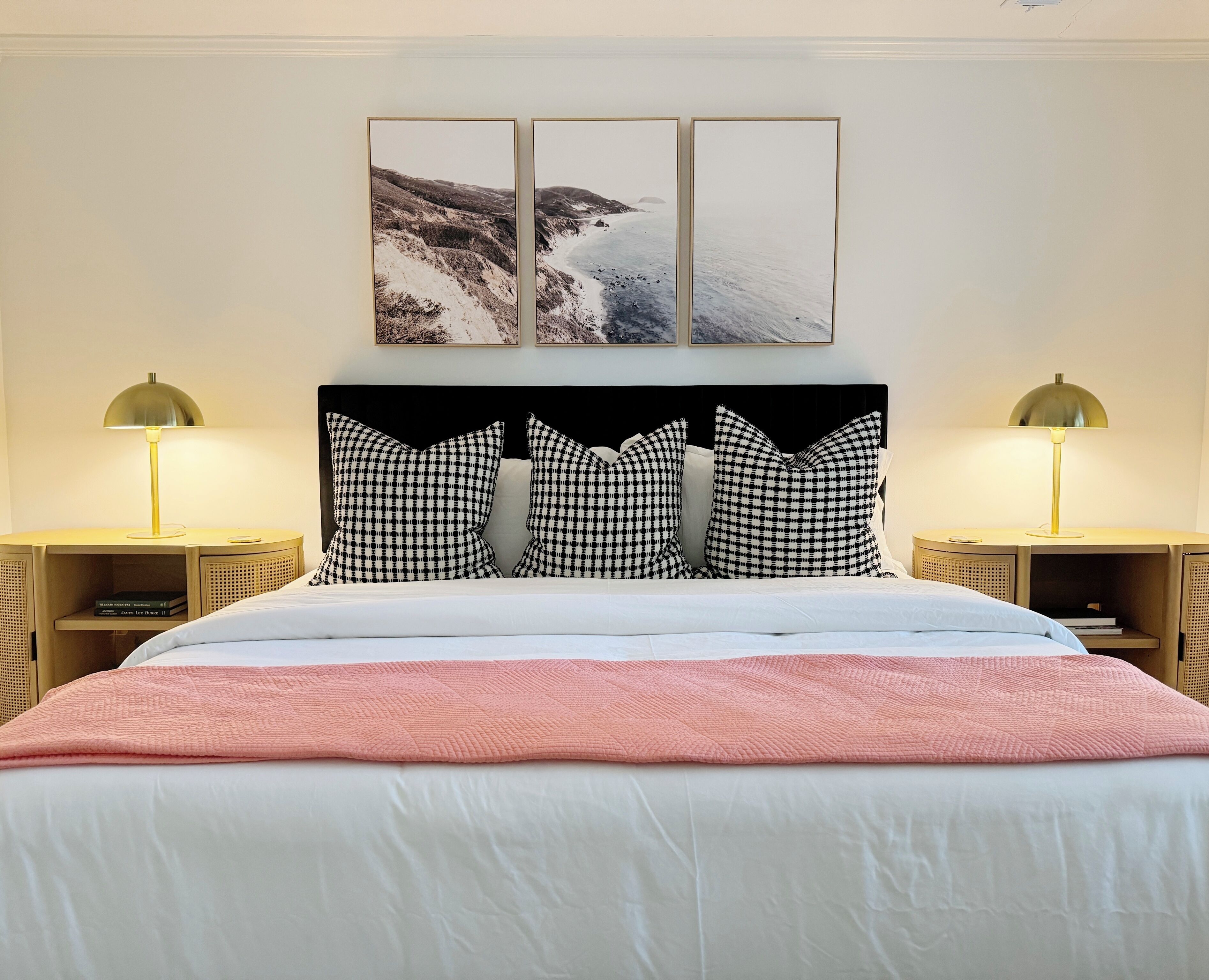 Ein ordentlich bezogenes Bett mit einer rosa Decke, karierten Kissen und einem Kopfteil mit einem Bild von einer Küstenlandschaft.