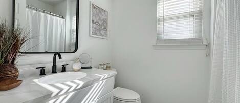 A bathroom with a white vanity, a black faucet, a white toilet, and a window with blinds.
