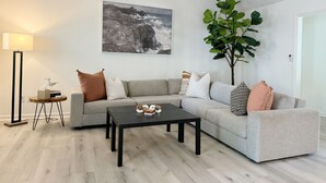 Ein modernes Wohnzimmer mit einem grauen Sofa, einem schwarzen Couchtisch, einer Stehlampe und einer großen Zimmerpflanze.