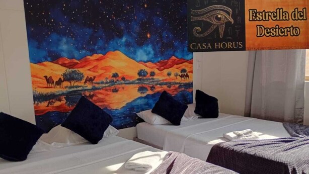 Une chambre d’hôtel avec deux lits, un tableau représentant une nuit étoilée accroché au mur, et une enseigne indiquant : « Estrella del Desierto CASA HORUS ».