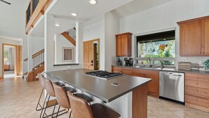 A modern kitchen with a central island, bar stools, wooden cabinets, stainless steel appliances, and a large window offering a view of greenery.