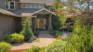 A two-story house with a front porch, a small garden with various plants, and a clear sky.