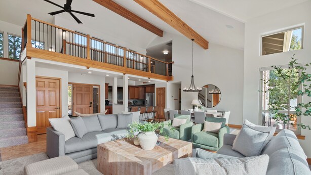 A spacious living area with a high ceiling, wooden beams, a staircase, and a view of the kitchen and dining area.