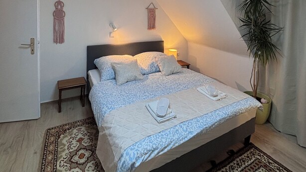 Ein Schlafzimmer mit Oberlicht, einem Bett mit gemustertem Bettbezug, einem Nachttisch, einer Stehlampe und einem Wandbild.