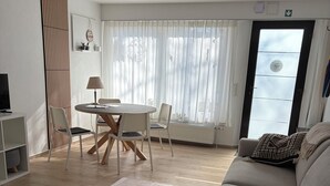 Ein modernes Wohnzimmer mit Essbereich, einem grauen Sofa, einem gemusterten Teppich und einem großen Fenster mit transparenten Vorhängen.