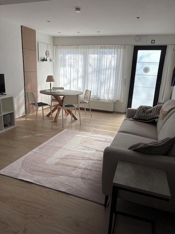 Ein modernes Wohnzimmer mit Essbereich, einem grauen Sofa, einem gemusterten Teppich und einem großen Fenster mit transparenten Vorhängen.
