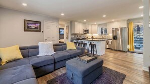A modern living room with a grey sectional sofa, a small ottoman, and a kitchen area with white cabinets and stainless steel appliances. - Private Woods - Heavenly California Lodge (South Lake Tahoe)