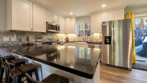 A modern kitchen with white cabinets, a large island with bar stools, stainless steel appliances, and a view of the outdoors. - Private Woods - Heavenly California Lodge (South Lake Tahoe)
