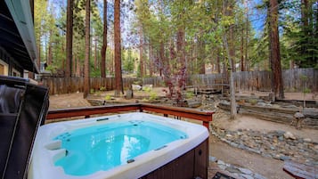 Un bain à remous dans un jardin à l’arrière d’une maison, entouré d’arbres, avec une terrasse en bois et un chemin en pierre.