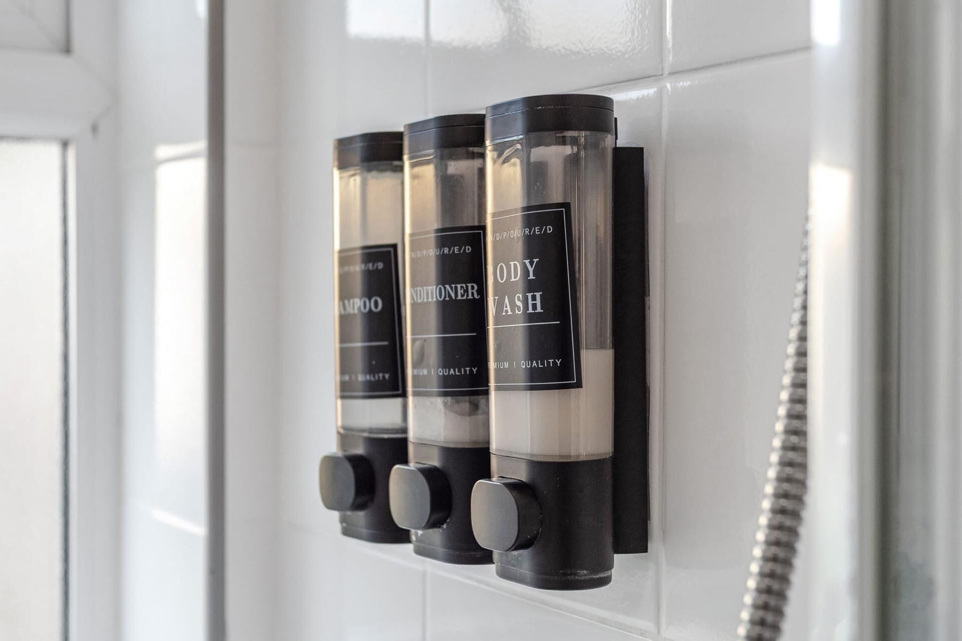 A bathroom with three black dispensers labeled 'BODY WASH,' 'SHAMPOO,' and 'SHAMPOO' mounted on a white tiled wall.