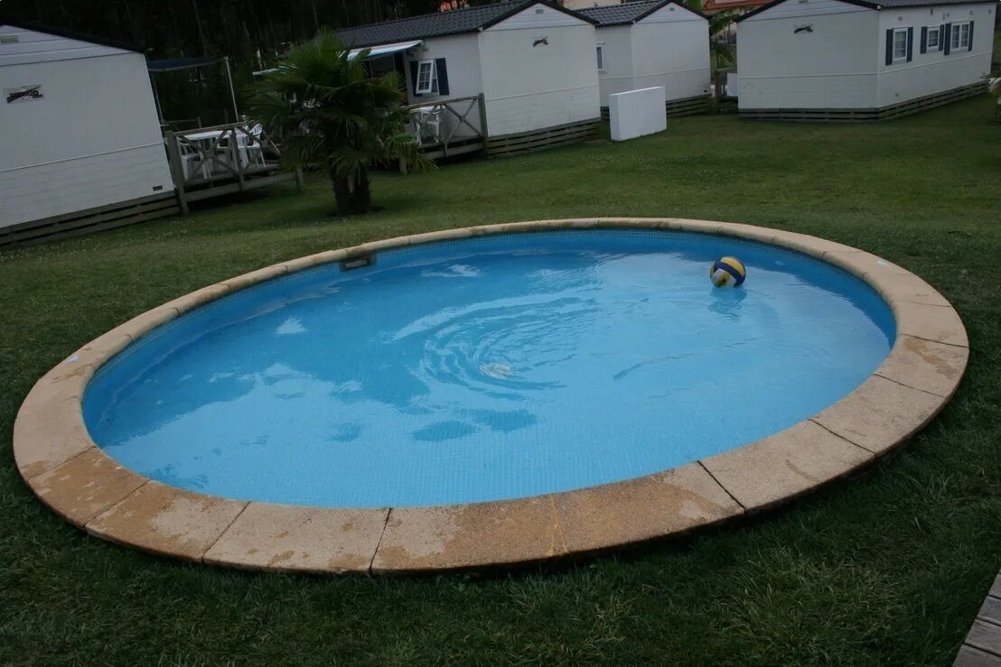 Ein runder Swimmingpool mit einer gefliesten Kante, umgeben von einer Rasenfläche und Mobilheimen im Hintergrund.