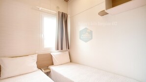 A small, clean room with two single beds, a window with curtains, and a wall-mounted shelf.