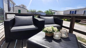 An outdoor seating area with two sofas and a table, a wooden deck, and a view of mobile homes in the background.
