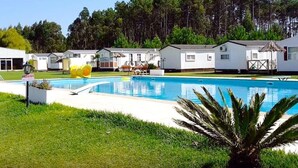 A swimming pool with a grassy area in front, surrounded by white cabins with a thatched roof.