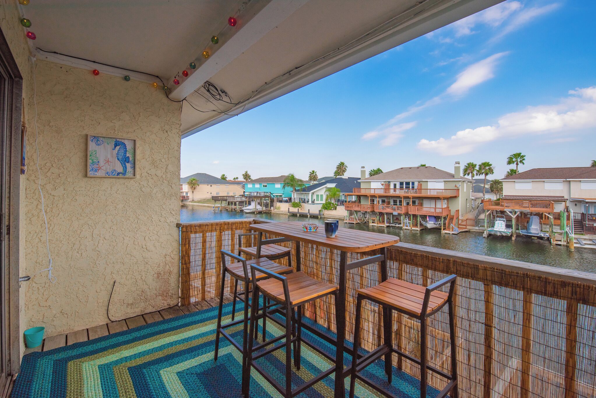 A balcony with wooden furniture, a view of a waterfront with buildings and boats, and a painting on the wall.