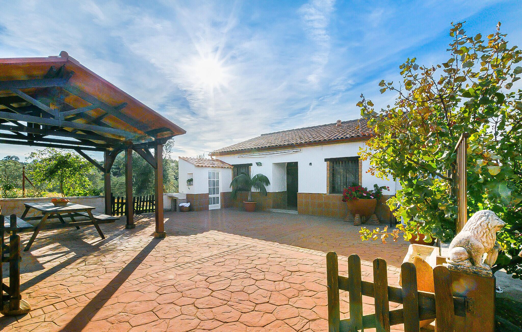 A covered patio area with a wooden table and chairs, a stone-tiled floor, and a white building with a tiled roof in the background.