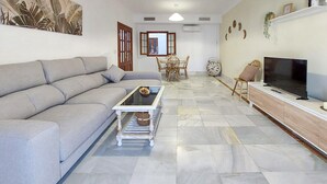 Un salon avec un canapé gris, une table basse en verre, un téléviseur posé sur un meuble en bois, et un coin repas avec une table et des chaises.