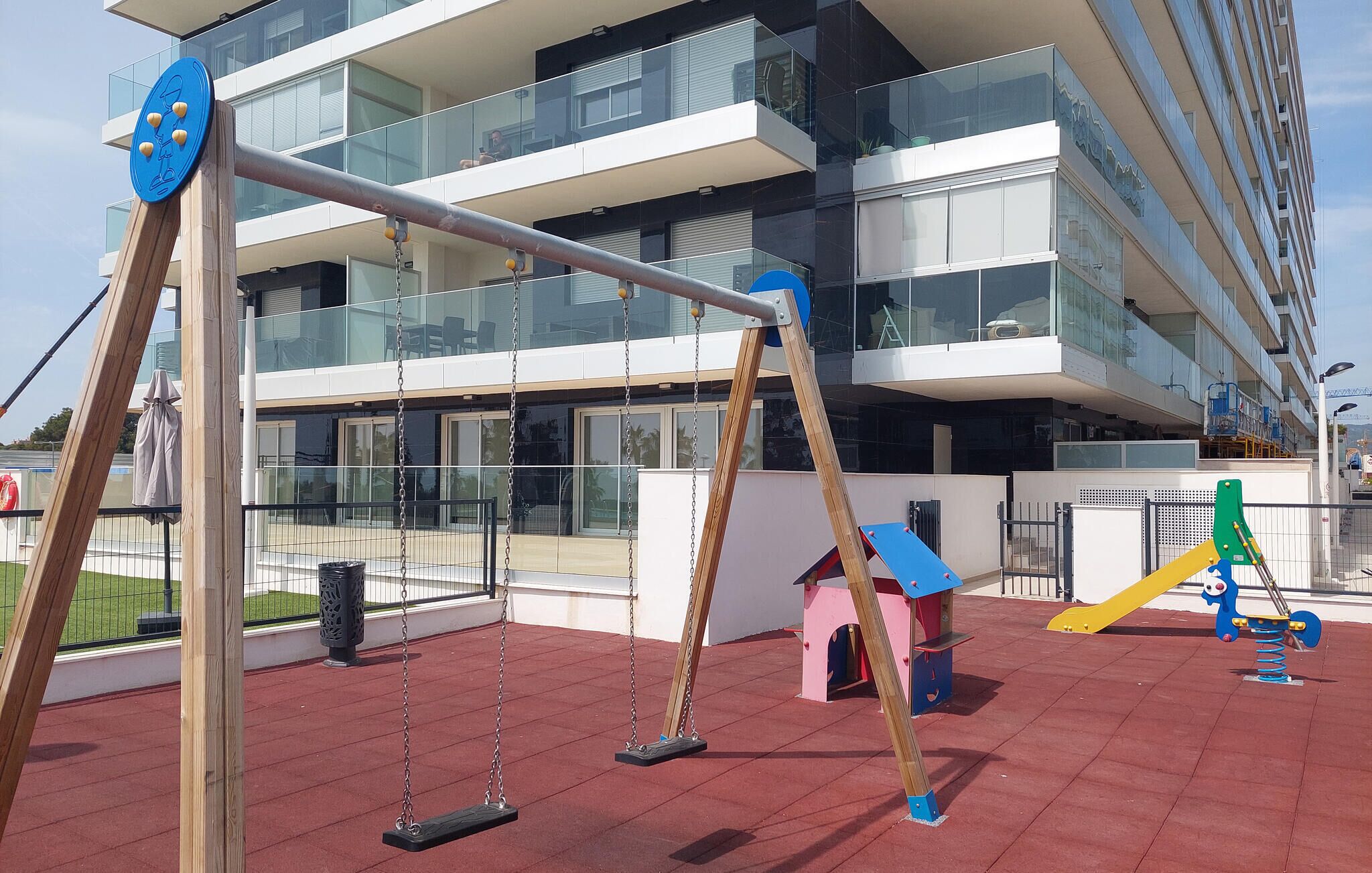 Un terrain de jeux avec des balançoires et une structure d’escalade, situé devant un bâtiment moderne doté de grandes fenêtres.