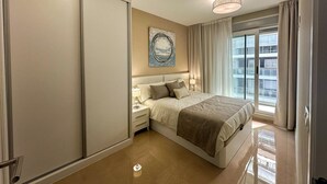 Habitación de hotel con cama, mesitas de noche, un cuadro en la pared, una ventana con cortinas y vistas a edificios.