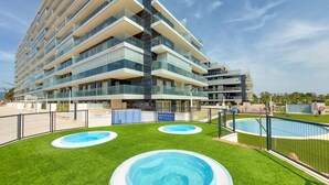 Un hotel de varios pisos con diseño moderno, que cuenta con balcones con barandas de vidrio, un área de piscina con césped artificial y un cielo azul despejado.