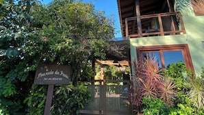 A two-story building with a wooden balcony, surrounded by lush greenery and a gravel pathway leading to a gated entrance. - Pousada da Praia Ferrugem (Garopaba)