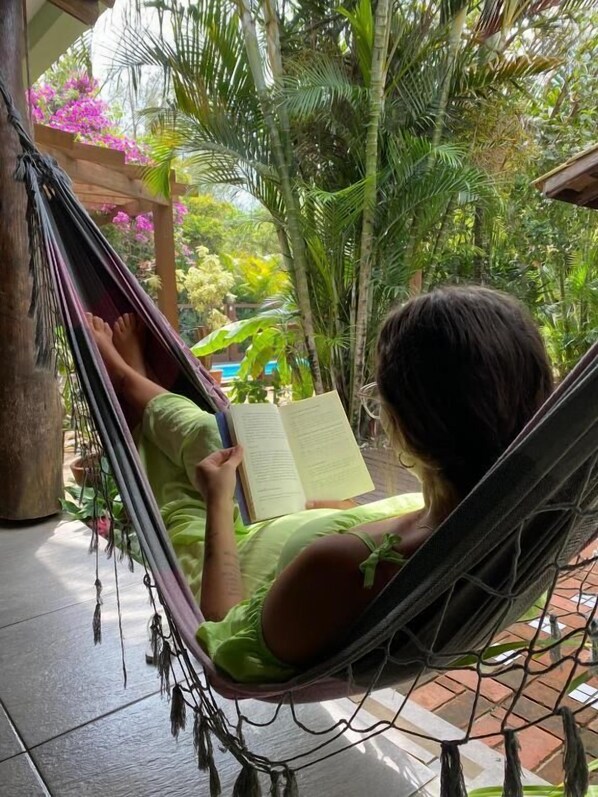 A person relaxing in a hammock on a porch with a book, surrounded by lush greenery and a pool. - Pousada da Praia Ferrugem (Garopaba)