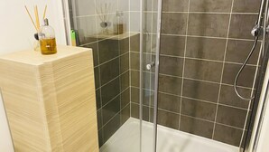 A modern bathroom with a glass-enclosed shower, dark tiled walls, and a wooden vanity with a bottle of liquid and reed diffusers.