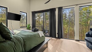 Une chambre dotée d’un lit, d’un téléviseur à écran plat, d’un ventilateur de plafond et de grandes fenêtres donnant sur la forêt.