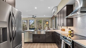 Une cuisine moderne dotée d’appareils électroménagers en acier inoxydable, de meubles de rangement aux couleurs sombres et offrant une vue sur les arbres grâce à de grandes fenêtres.