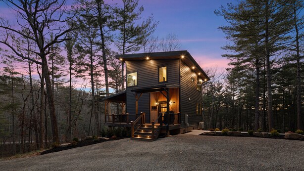 Une cabane à deux étages, avec une façade en bois, un porche à l’avant et une allée de gravier entourée d’arbres.