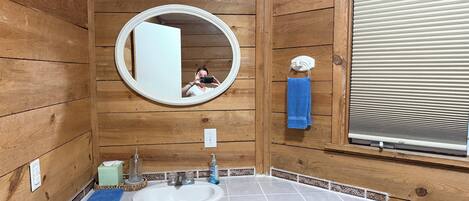 A wooden cabin-style bathroom with a round mirror, a sink, and a window with blinds.