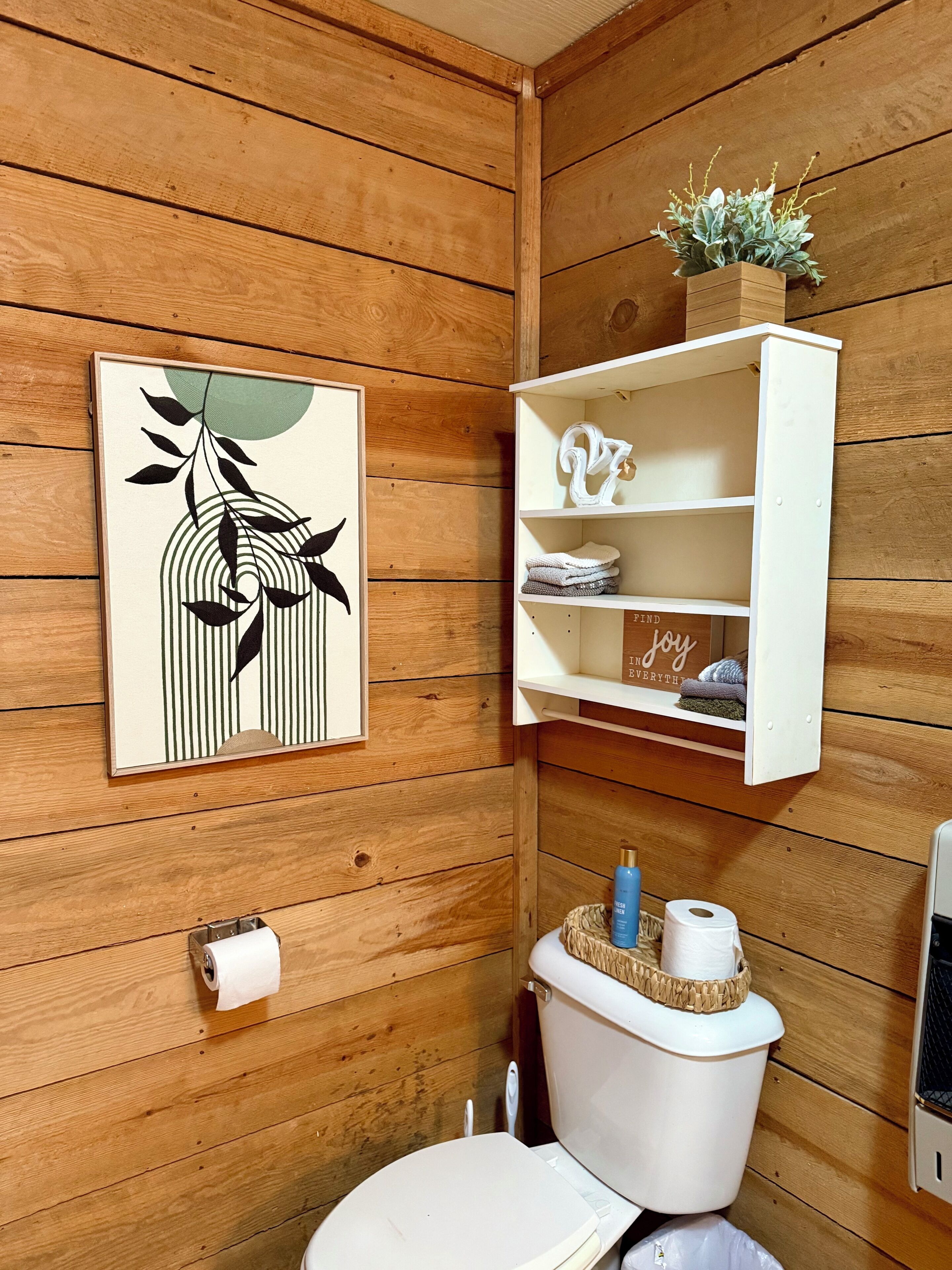 A wooden-paneled bathroom with a toilet, a framed abstract artwork, a white shelf with towels and a decorative item, and a small plant on the shelf.