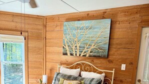 A bedroom with a wooden paneled wall, a bed with a plaid bedspread, a painting of trees, and a window with a view of trees. - Eagle’s Nest Cabin • Private Forest Stay (Caddo Gap)