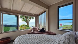 A bedroom with a large bed, two windows with ocean views, a stop sign outside, and a chair by the window. - Justavacation Beachfront Bach (Riverton / Aparima)