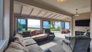A spacious living room with a large sofa, armchairs, and a coffee table. There is a TV mounted on the wall and a ceiling fan. The room has large windows offering a view of the outdoors. - Justavacation Beachfront Bach (Riverton / Aparima)