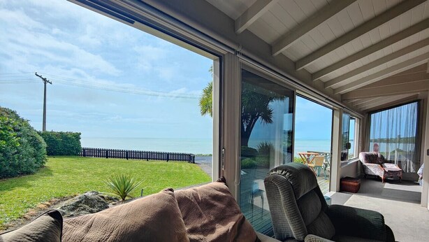 A modern living room with a large glass door leading to a balcony overlooking a beach. - Justavacation Beachfront Bach (Riverton / Aparima)
