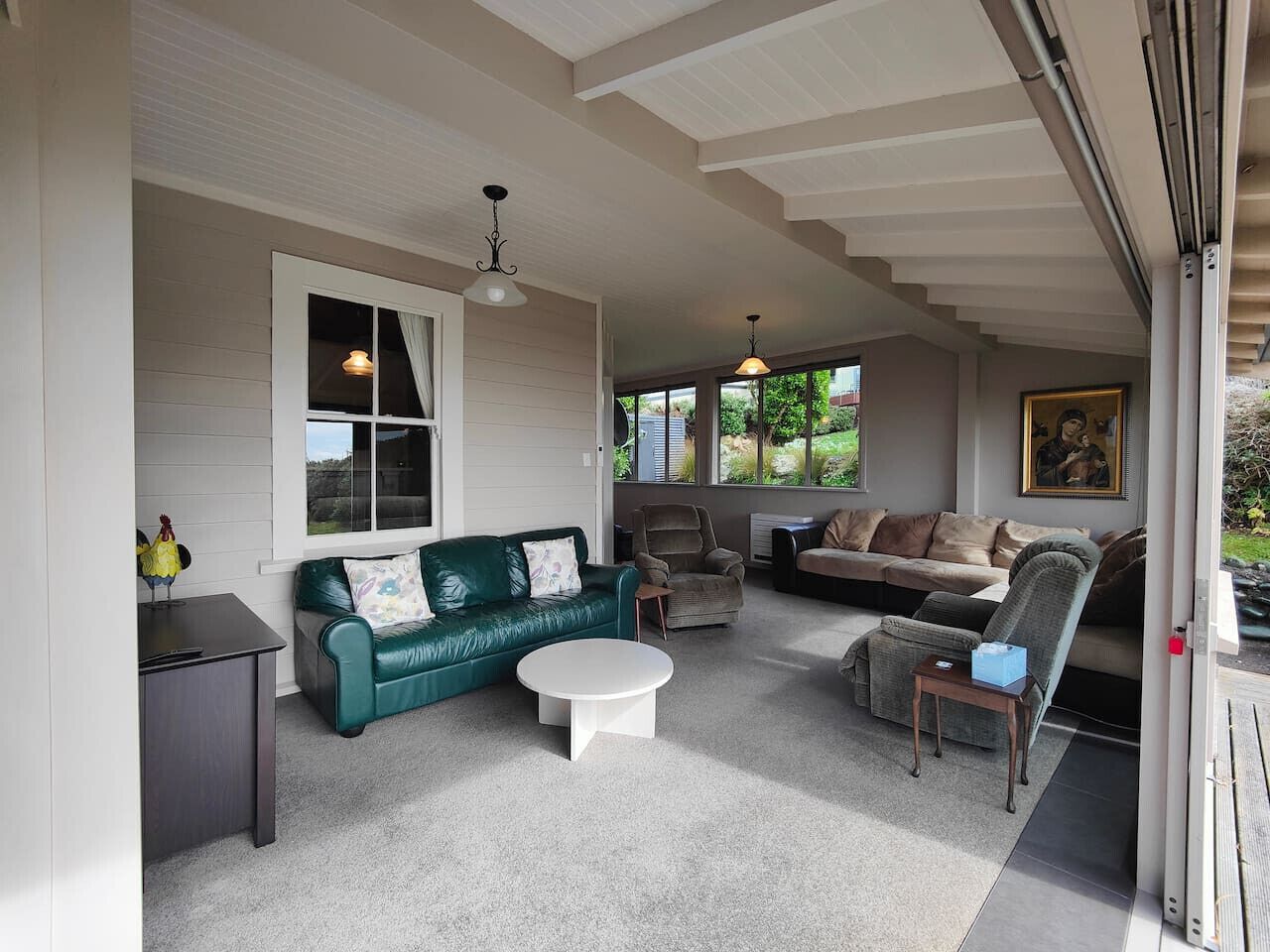 A spacious living room with a green leather sofa, a white coffee table, and a grey armchair. There is a window with a view of greenery outside.