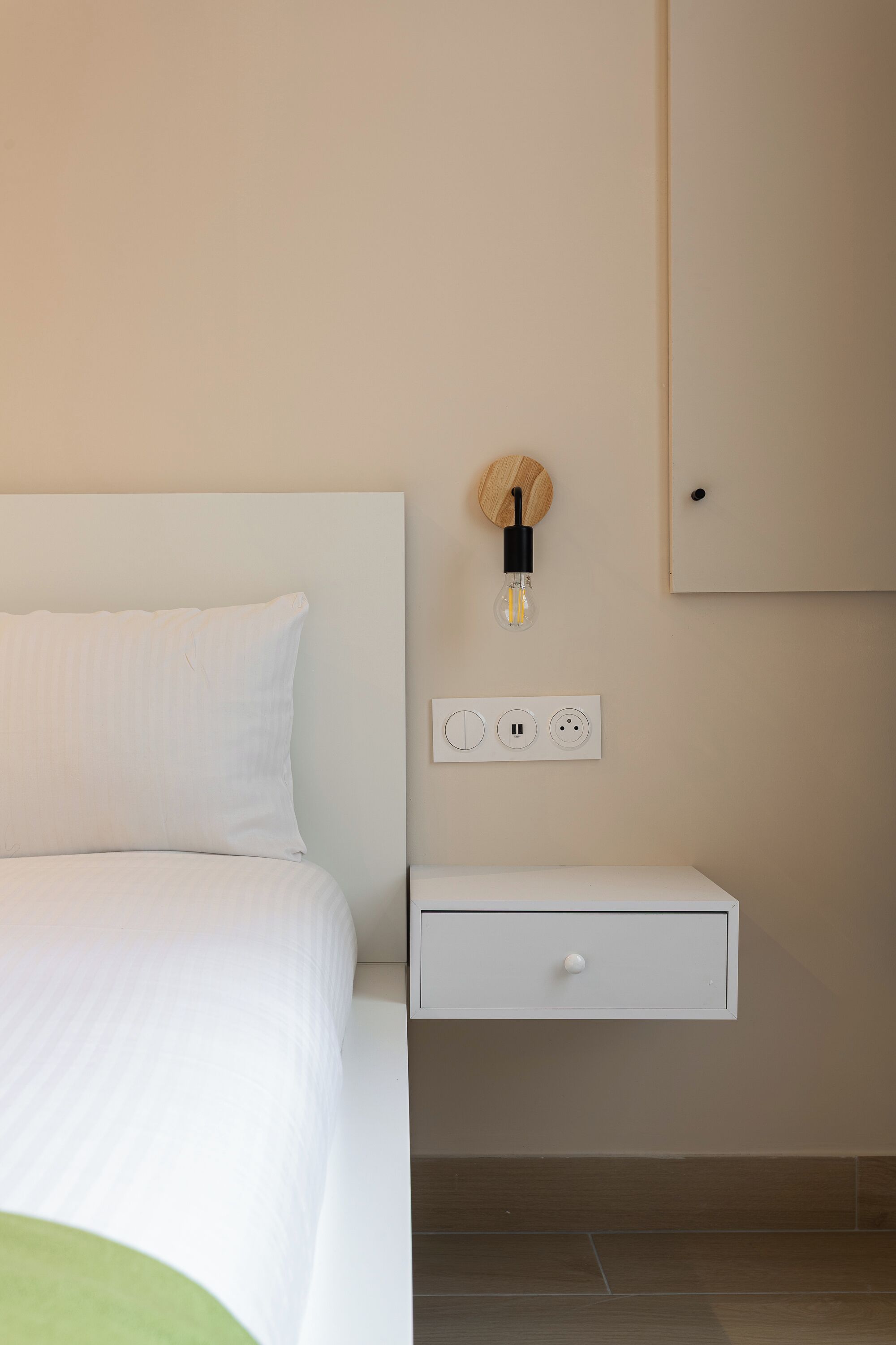 Ein modernes Hotelzimmer mit einem ordentlich bezogenen Bett, einem Nachttisch mit Lampe und einer wandmontierten Steckdose.
