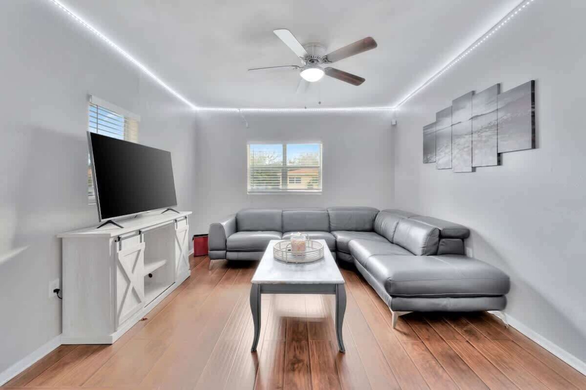 A modern living room with a grey sectional sofa, a flat-screen TV on a white cabinet, a ceiling fan, and a window with blinds.