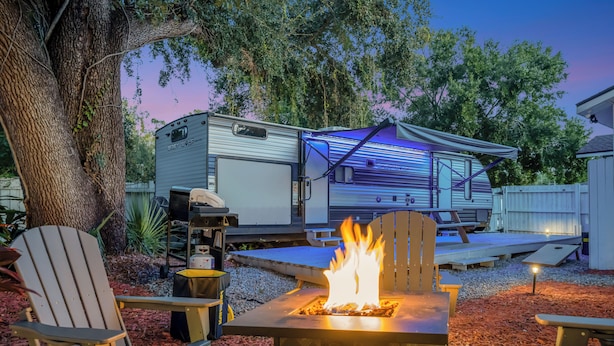 Un terrain de camping équipé d’un foyer, de chaises Adirondack et d’une maison mobile en arrière-plan.