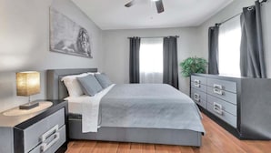 Une chambre à coucher moderne avec un lit, une table de chevet, une commode, un ventilateur de plafond et un tableau au mur.