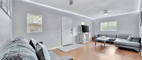 A spacious living room with a grey sofa, a coffee table, and a TV on a white cabinet. There are two ceiling fans and a door leading to another room.
