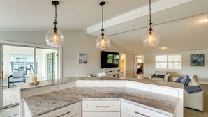 Una cocina moderna con una isla grande, armarios blancos y encimera de piedra. Hay lámparas colgantes del techo.