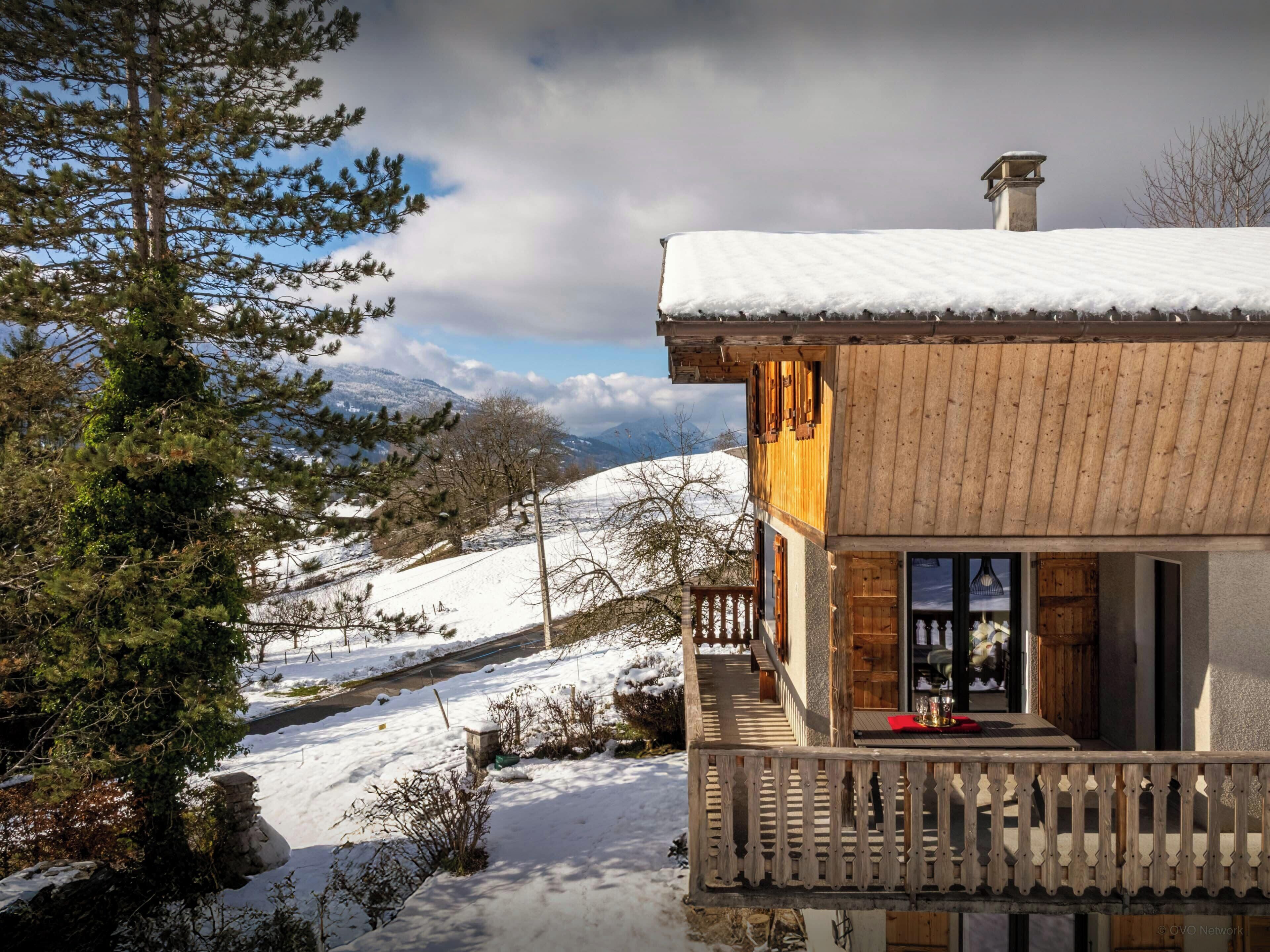 A wooden chalet with a balcony overlooking a snowy landscape and mountains.