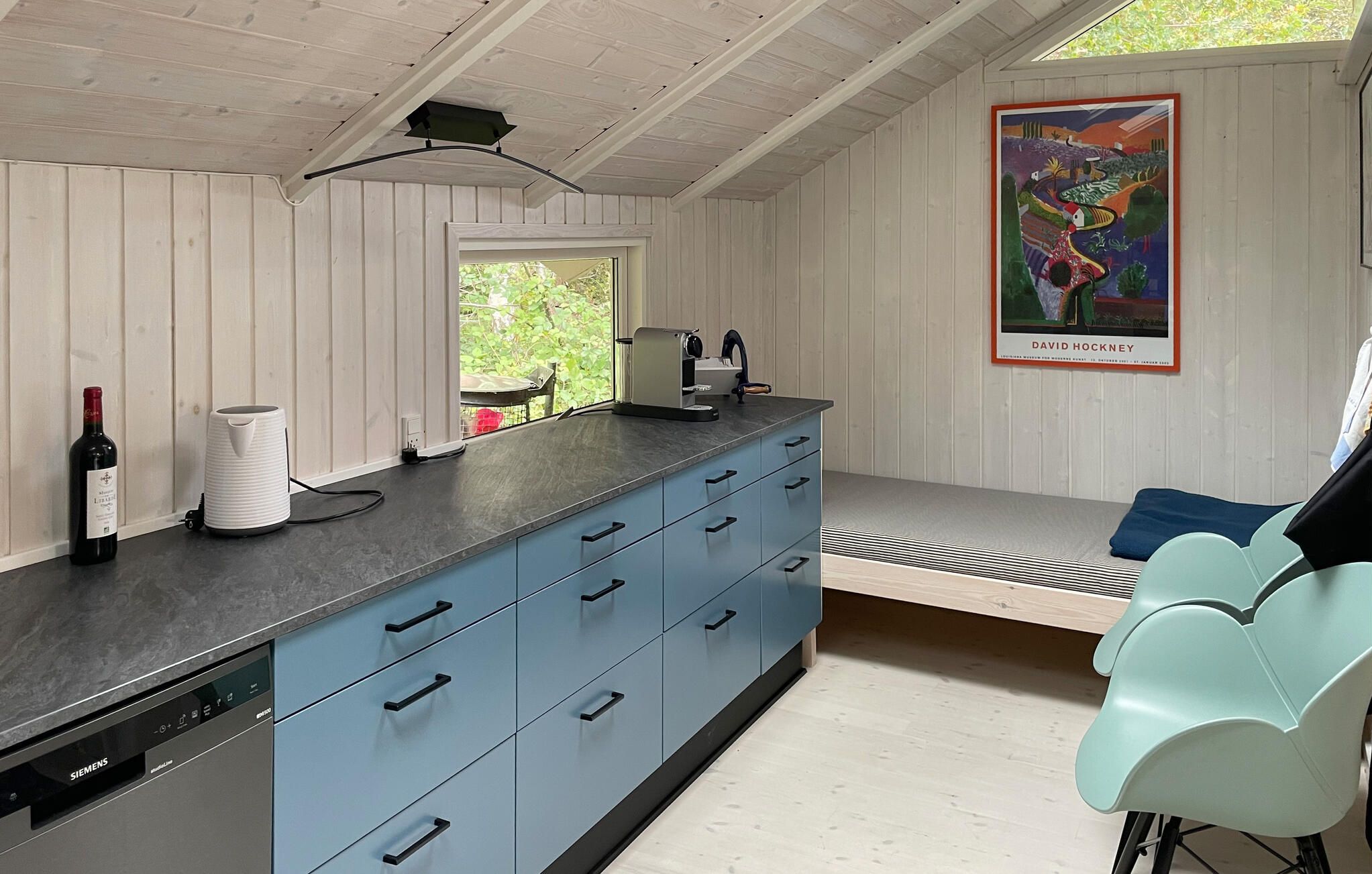A modern kitchen with a Siemens dishwasher, a coffee machine, and a wine bottle. There's a window with a view of greenery, a painting by David Hockney, and a teal chair.